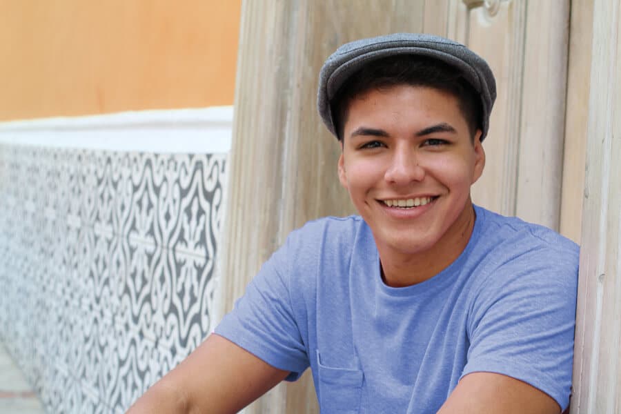 A smiling teenage boy wearing a flat cap is leaning against a doorframe with a patterned tile background, symbolizing hope and positivity in his Charlotte drug treatment program.
